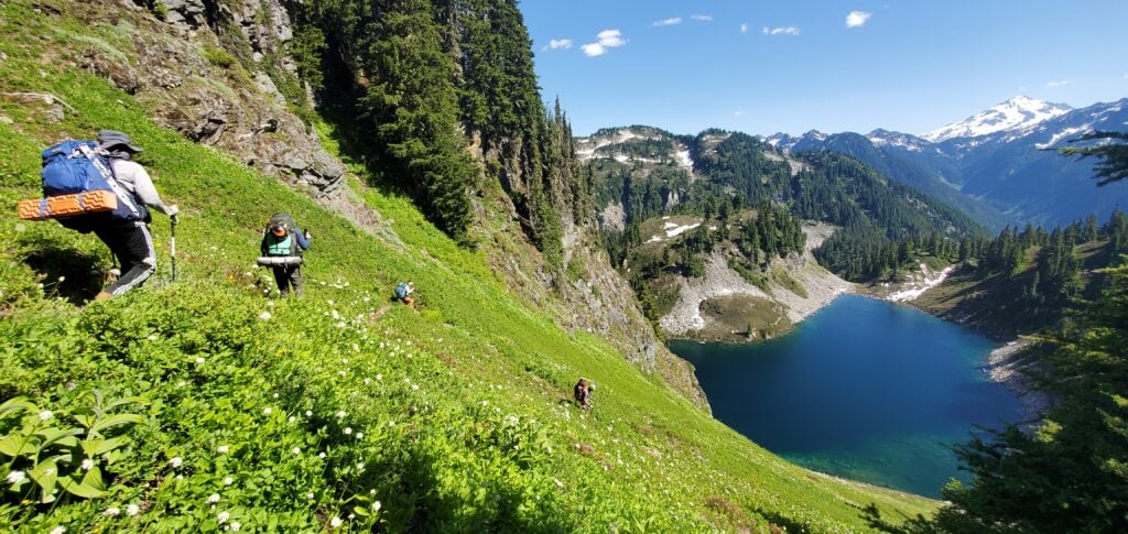 Climbers hiking down a lush green hillside towards a mountain lake.