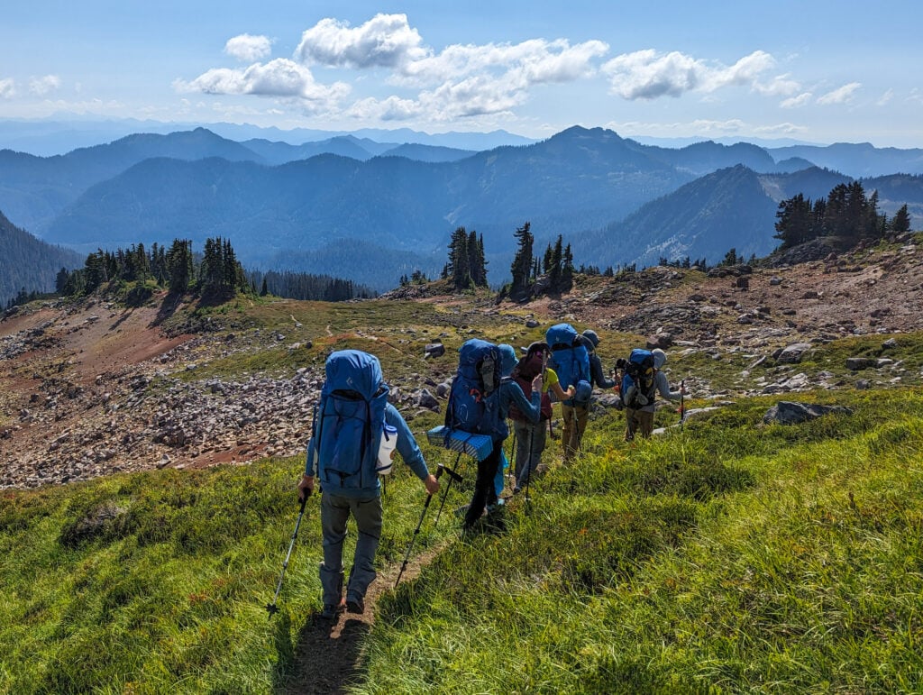 A line of backpackers trek through an alpine meadow on a well-worn trail in the backcountry of the North Cascades.