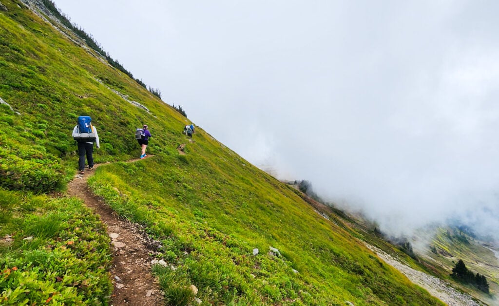 Backpackers walk along an alpine meadow towards the fog of a Cascade Mountain pass in Washington
