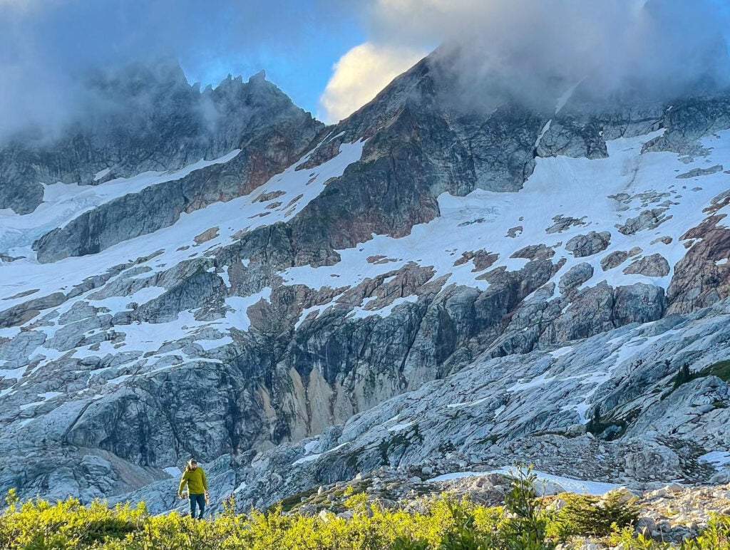 A camper swallowed by the large mountains that loom in the background of the North Cascades alpine wilderness on an American Alpine Institute backpacking trip
